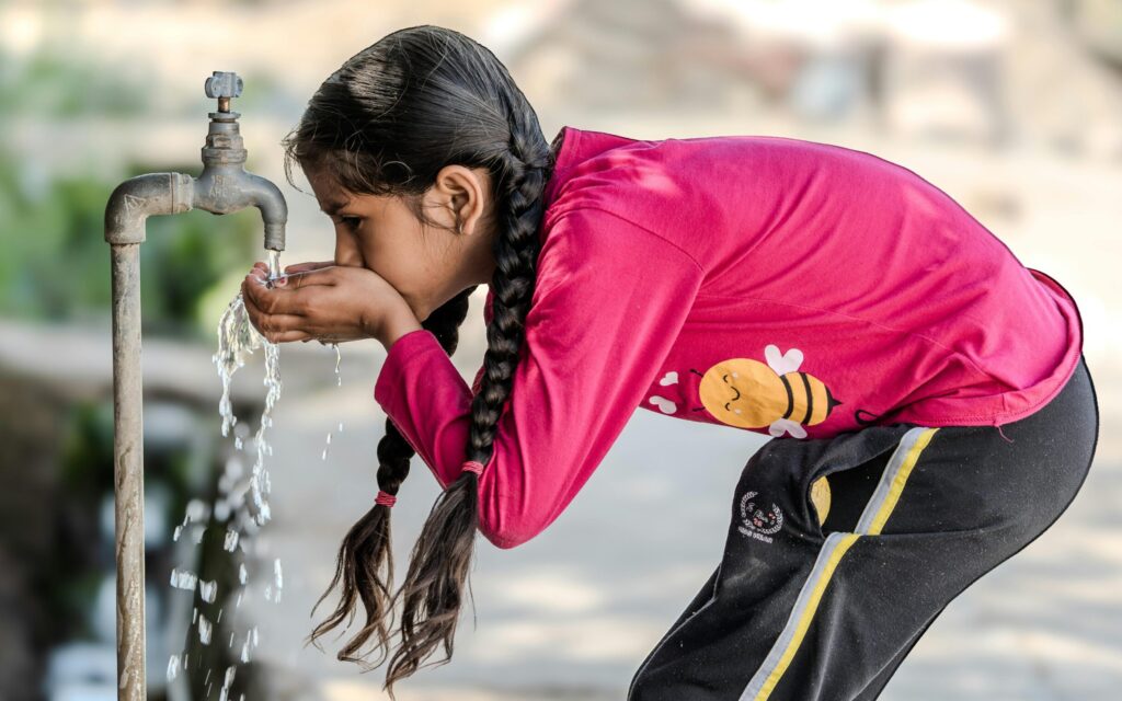 A young girl drinks water from an outdoor tap on a sunny day, emphasizing the simplicity of rural life.