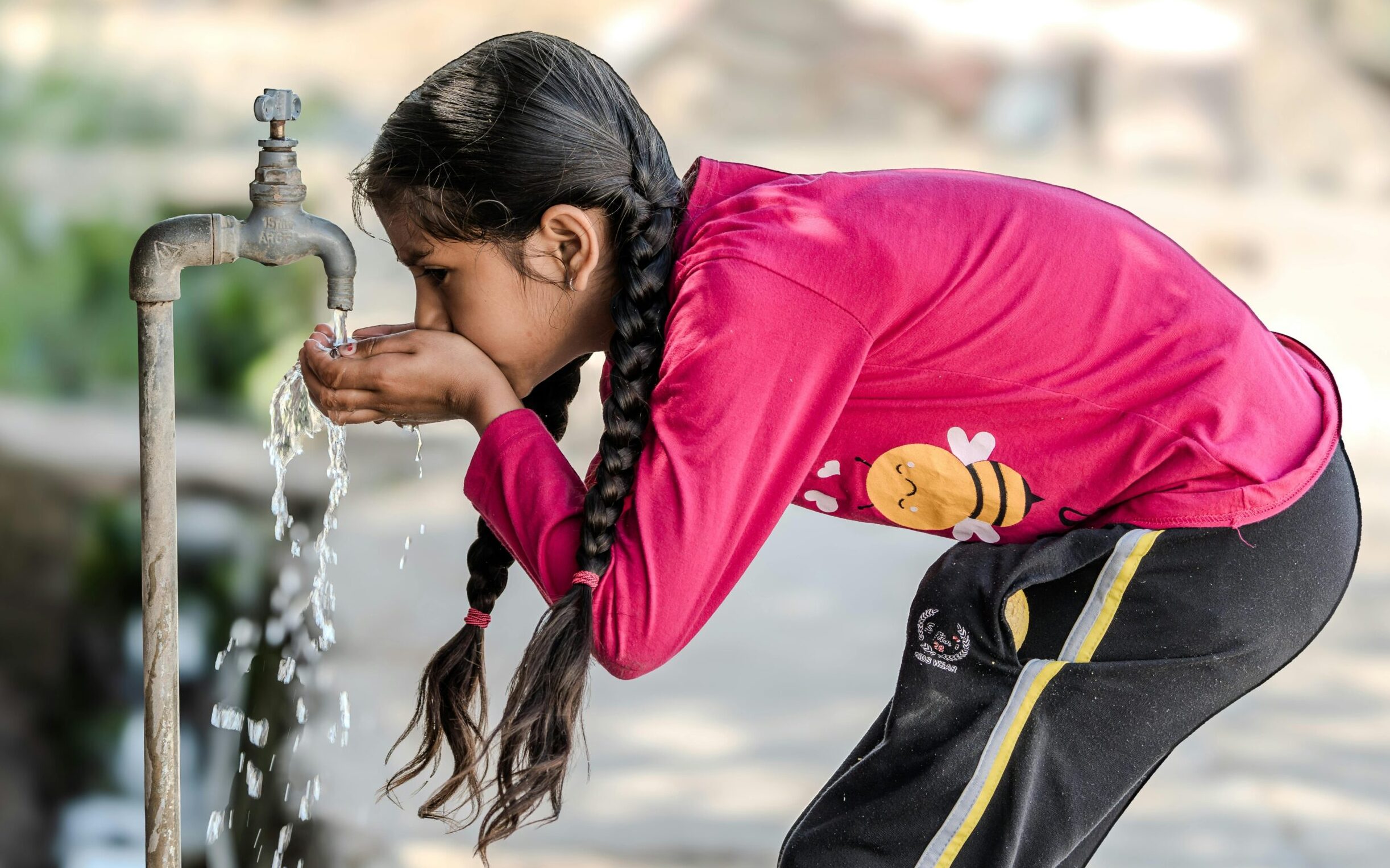 A young girl drinks water from an outdoor tap on a sunny day, emphasizing the simplicity of rural life.
