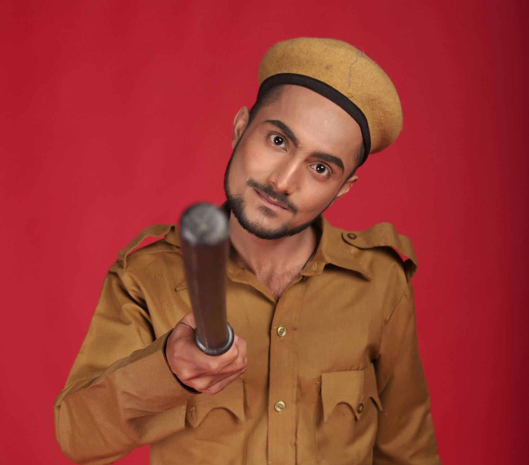 Portrait of an Indian policeman holding a baton against a red backdrop in a studio.